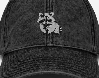 May include: A black, vintage-style baseball cap with a distressed finish. The cap features a white graphic of a raccoon on the front. The cap has a curved brim and an adjustable strap at the back.