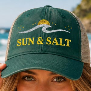 May include: A dark green baseball cap with a mesh back, featuring a graphic of a silver wave and a yellow sun above the text SUN & SALT in yellow lettering. The cap has a structured front panel and a curved brim, designed with a casual aesthetic.