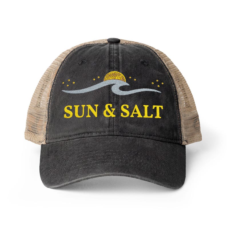 May include: A black baseball cap with a tan mesh back. The front features an embroidered design of a yellow sun, a light gray wave, and small yellow stars. Below the graphic, the text SUN & SALT is embroidered in yellow capital letters.