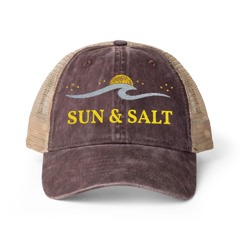 May include: A dark brown distressed trucker hat with a mesh back. The front panel features an embroidered design of a yellow sun, a light blue wave, and small yellow stars. The text SUN & SALT is embroidered in yellow below the graphic.