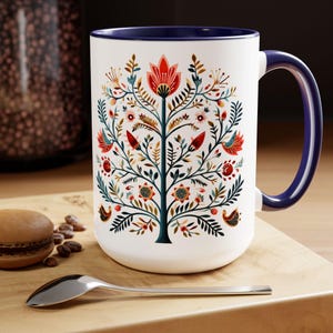 May include: White ceramic mug with a blue rim and handle. The mug features a colorful folk art style tree design with red, green, blue, yellow, and orange flowers and birds.