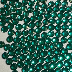 May include: A large quantity of emerald green rhinestones, also known as crystals or gems, are scattered on a white surface. The rhinestones are faceted and have a shiny, reflective finish.