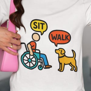 May include: White t-shirt with a cartoon illustration of a person in a wheelchair and a yellow dog. Speech bubbles say "SIT" and "WALK". The person is wearing blue pants and the dog is standing.