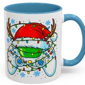 May include: White ceramic mug with a blue interior and handle. The mug features a festive design of a video game controller wearing a Santa hat and reindeer antlers, wrapped in Christmas lights and snowflakes.
