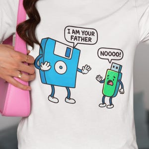 May include: White t-shirt featuring a cartoon illustration of a floppy disk and a USB drive. The floppy disk says "I AM YOUR FATHER" and the USB drive says "NOOOO!". The shirt is being worn by a person.