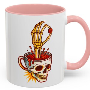 May include: White ceramic mug with a pink interior and handle. The mug features a skull design with a skeleton hand holding a coffee bean. The design is in red, yellow, and brown.