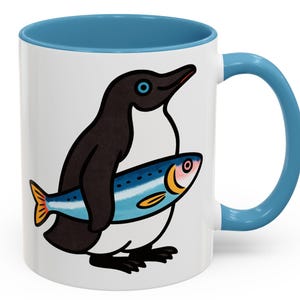 Peut inclure: Mug en céramique blanche avec intérieur et anse bleus. Le mug présente un dessin animé d'un pingouin tenant un poisson. Le pingouin est noir et blanc, et le poisson est bleu avec des accents orange et jaune.
