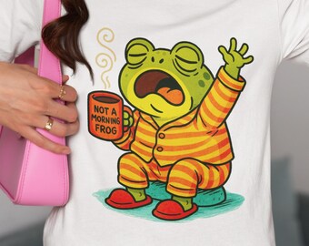 May include: White t-shirt featuring a cartoon frog in orange and yellow striped pajamas, yawning and holding a mug that reads "NOT A MORNING FROG." The frog is sitting on a blue cushion with red slippers.