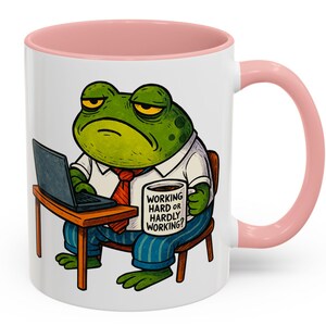 May include: A white ceramic mug with a pink interior and handle. The mug features a cartoon frog wearing a shirt and tie, working on a laptop. The frog is holding a mug that says "WORKING HARD OR HARDLY WORKING?"