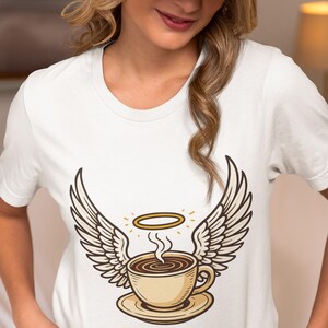 May include: White t-shirt featuring a coffee cup with angel wings and a halo. The coffee cup is beige with steam rising, and the wings are detailed with brown outlines. The design is centered on the shirt.
