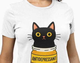 May include: White t-shirt featuring a cartoon black cat peeking out of a yellow pill bottle labeled "ANTIDEPRESSANT." The cat has yellow eyes and whiskers. Three yellow pills are scattered below the bottle.
