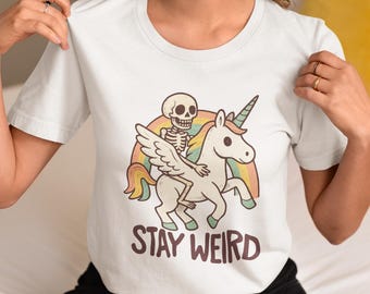 May include: White t-shirt featuring a cartoon skeleton riding a unicorn with rainbow wings and a rainbow background. The text "STAY WEIRD" is printed below the graphic. The shirt has short sleeves.