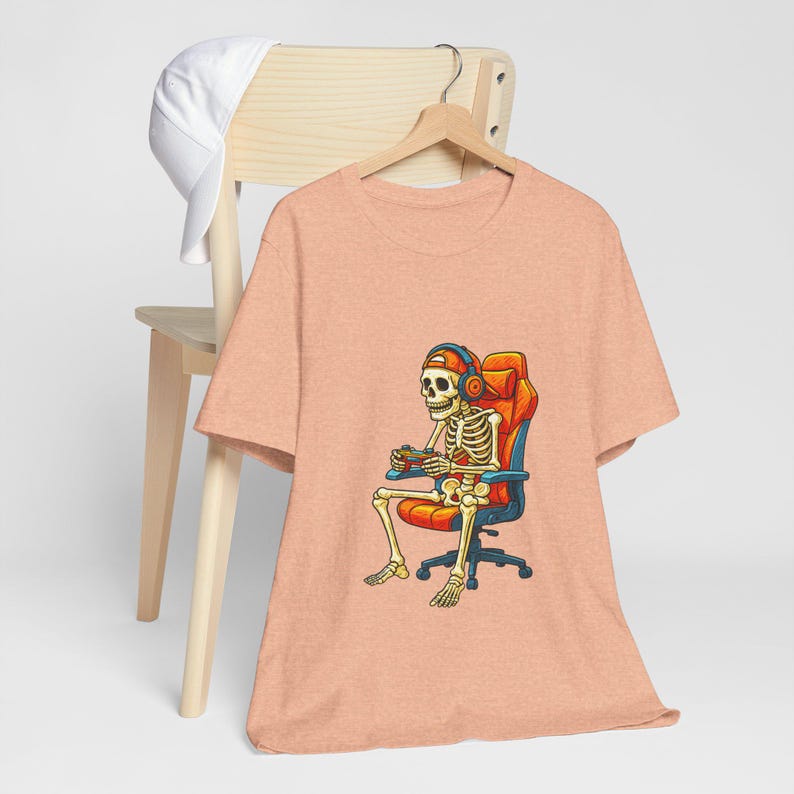 May include: Peach-colored t-shirt featuring a skeleton gamer wearing headphones and a cap, seated in an orange and blue gaming chair, holding a controller. A white cap rests on a wooden chair in the background.