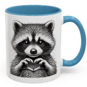 May include: White ceramic coffee mug with a blue rim and handle. The mug features a black and white illustration of a raccoon making a heart shape with its paws.