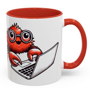 May include: A white ceramic coffee mug with a red rim and handle. The mug features a cartoon illustration of a red crab wearing glasses and using a laptop computer.