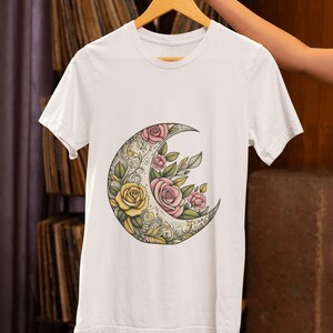 May include: White t-shirt featuring a crescent moon design filled with yellow and pink roses and green leaves. The floral illustration is detailed with ornate patterns. The shirt is hanging on a wooden hanger.
