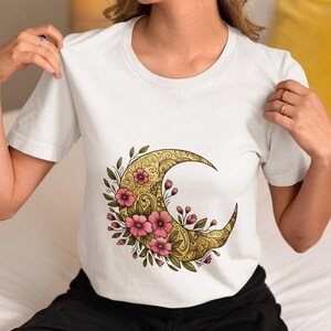 May include: White t-shirt featuring a golden crescent moon design with intricate patterns and pink floral accents. The design is centered on the front of the shirt. The shirt has short sleeves and a crew neck.