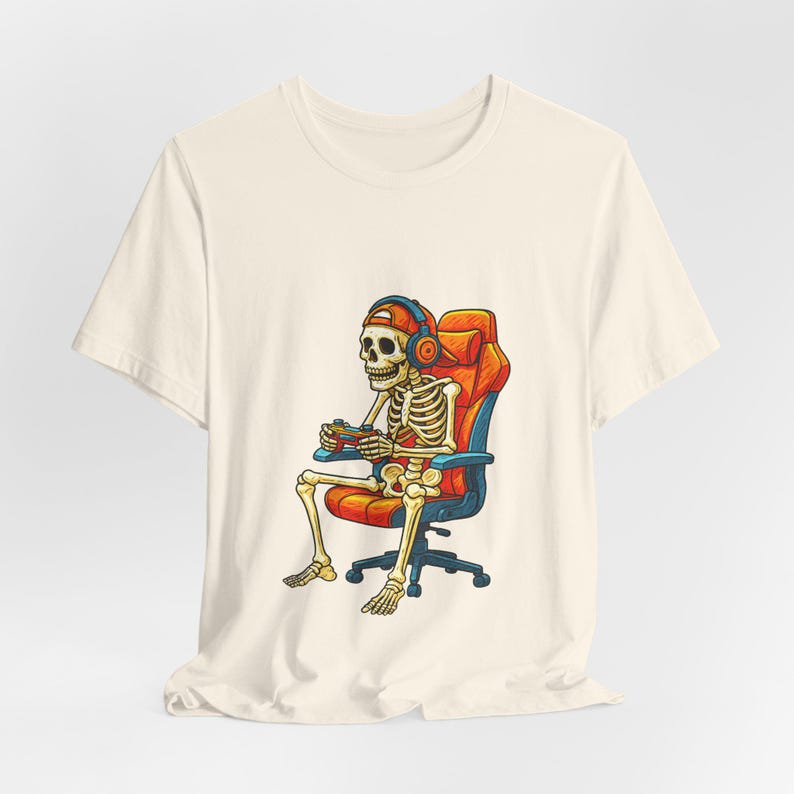May include: Cream-colored t-shirt featuring a cartoon skeleton gamer. The skeleton is seated in an orange and blue gaming chair, wearing headphones and a cap, and holding a game controller. The design has a retro, vintage style.