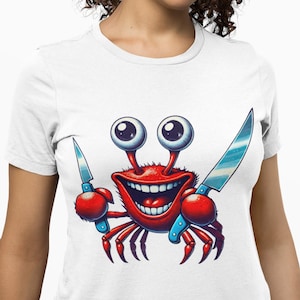 May include: White t-shirt featuring a cartoon crab with large, googly eyes and a wide grin. The crab is red with blue accents and holds two knives. The design is a playful and humorous illustration.