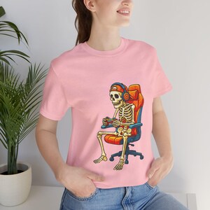 Cartoon skeleton wearing a backwards cap and headphones, sitting in an orange gaming chair while holding a game controller. High-contrast, playful gamer artwork for a unisex T-shirt in adult, youth, and toddler sizes—great for Halloween, streaming, LAN parties, and gifts for console fans.