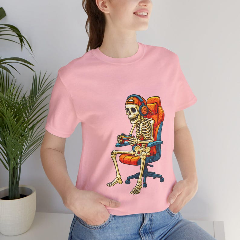 Cartoon skeleton wearing a backwards cap and headphones, sitting in an orange gaming chair while holding a game controller. High-contrast, playful gamer artwork for a unisex T-shirt in adult, youth, and toddler sizes—great for Halloween, streaming, LAN parties, and gifts for console fans.