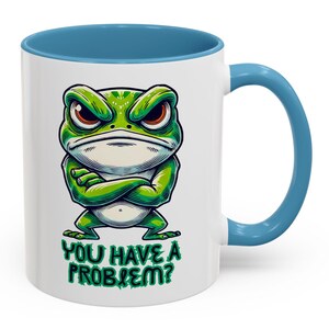 May include: White ceramic mug with a blue rim and handle. The mug features a cartoon illustration of a green frog with crossed arms and an angry expression. The text "YOU HAVE A PROBLEM?" is printed below the frog.