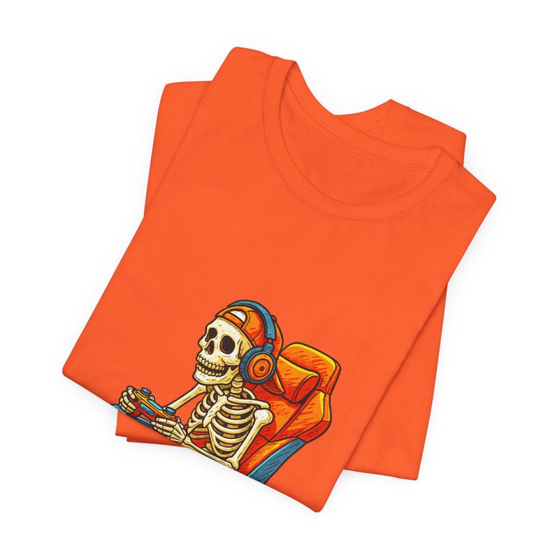 May include: Orange t-shirt featuring a skeleton gamer wearing headphones and holding a game controller. The skeleton is seated in a gaming chair. The design is in a cartoon style.