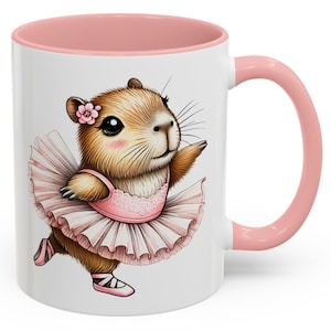 May include: A white ceramic mug with a pink rim and handle. The mug features a cartoon illustration of a brown capybara wearing a pink tutu and pink ballet shoes. The capybara has a pink flower in its hair.
