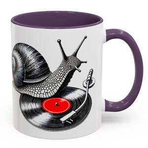 May include: A white ceramic coffee mug with a purple rim and handle. The mug features a black and white illustration of a snail playing a record on a turntable. The snail has a shell with a black and white pattern. The record is black with a red center.