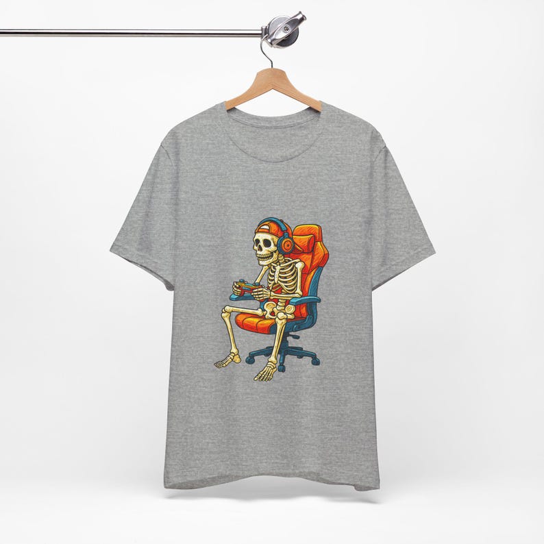 May include: A gray t-shirt featuring a cartoon skeleton wearing headphones and playing a video game while seated in an orange and blue gaming chair. The skeleton is holding a game controller. The shirt is hanging on a wooden hanger.
