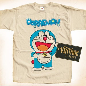 May include: A beige t-shirt with a vintage design featuring the cartoon character Doraemon. The design includes the character's name in a colorful font and a picture of Doraemon holding a sign that says "Vintage T-Shirt".