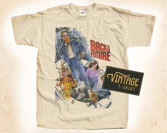 Könnte beinhalten: Ein Vintage-T-Shirt in Beige mit einem Grafikdruck des Films "Zurück in die Zukunft". Der Druck zeigt die Hauptfiguren des Films, darunter Marty McFly, Doc Brown und Jennifer Parker. Der Text "Zurück in die Zukunft" ist in roten und schwarzen Buchstaben gedruckt.