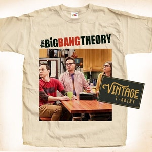 Puede incluir: Una camiseta beige de aspecto vintage con una foto del reparto de la serie de televisión The Big Bang Theory. El texto "the BIG BANG THEORY" está impreso en letras rojas y negras sobre la foto. El texto "Vintage T-Shirt" está impreso en letras negras sobre un rectángulo marrón en la esquina inferior derecha de la imagen.