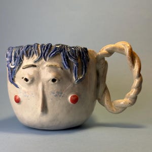 Könnte beinhalten: Eine skurrile Keramik-Tasse mit einem Gesichtsdesign. Die Tasse hat blaue Haare, rote Wangen und einen gedrehten Henkel. Die Tasse ist weiß mit blauen und roten Akzenten, ideal für Kaffee oder Tee.