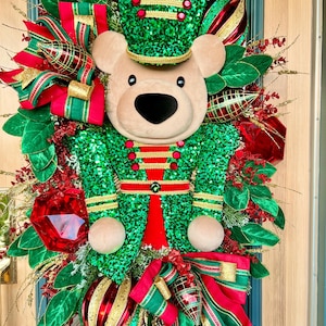 May include: A decorative wreath featuring a plush bear head wearing a green sequined nutcracker hat and uniform. The wreath is adorned with red and green ribbons, shiny red and gold ornaments, green fabric leaves, and gold glittered accents.