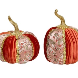 May include: Two decorative pumpkins with gold stems. The pumpkins feature a combination of patterned fabric and pleated orange velvet. Gold trim with sequins accents the fabric panels. These fall-themed decorations are perfect for autumn.