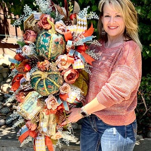 May include: A fall-themed decorative arrangement featuring two ornate teal pumpkins with gold accents, surrounded by peach-colored roses, orange ribbons, and decorative elements. The arrangement is held by a person.