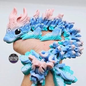 May include: A 3D-printed articulated dragon toy in shades of blue and pink. The dragon has a detailed design with scales, horns, and a long, flexible body. The toy is held in a hand, showcasing its size and flexibility.