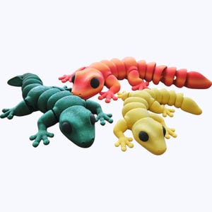 May include: Three brightly coloured, articulated lizard sculptures in green, orange, and yellow. Each lizard has a segmented body, four legs, and large black eyes. The orange lizard is at the top, the green lizard is on the left, and the yellow lizard is on the right.