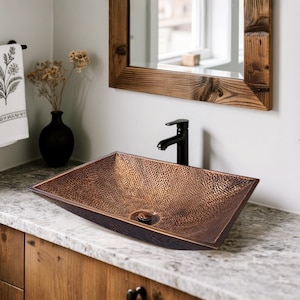 May include: A rectangular copper sink with a hammered texture sits on a marble countertop. The sink has a dark brown rim and a central drain. A black faucet is mounted on the wall behind the sink. The sink is part of a bathroom fixture.