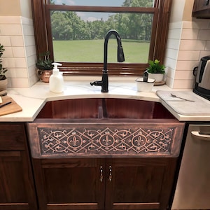 May include: A copper farmhouse sink with a decorative front panel featuring an ornate fleur-de-lis design. The sink is installed in a kitchen with a dark wood cabinet base and a black faucet. The background includes a window overlooking a green field.
