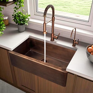May include: A copper-toned farmhouse sink with a matching faucet and a separate water dispenser. The faucet has a coiled spring design and is positioned over the sink, with water flowing into the basin. The sink is set into a wooden cabinet.