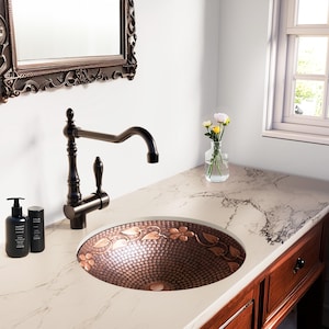 May include: A bathroom sink with a copper basin featuring a floral design. The sink is set into a white countertop with a dark bronze faucet. A mirror hangs on the wall above the sink, and a window is visible in the background.