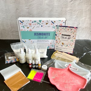 May include: A Jesmonite Starter Kit with a variety of materials for creating resin crafts. The kit includes a bag of white powder, three bottles of clear liquid, a small jar of sealer, a bag of colorful chips, two sheets of gold leaf, four bottles of liquid color, two sheets of neon pigment, and a pink ginkgo leaf mold.