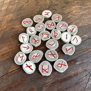 May include: A collection of rune stones, each with a unique red symbol, arranged on a rustic wooden surface. The stones are circular and appear to be made from a light-coloured material, possibly bone or antler.