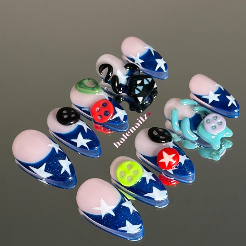 May include: A set of ten press-on nails featuring a blue base with white star patterns and a nude-colored cuticle area. Several nails are adorned with three-dimensional elements including black, red, and lime green buttons and a black cat figure.