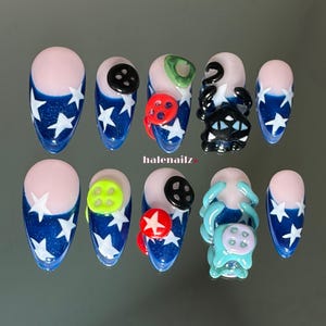 May include: A set of ten press-on nails featuring a dark blue base with white star patterns and a nude-colored cuticle area. The nails are decorated with various three-dimensional elements including black, red, and neon green buttons and cat figures.