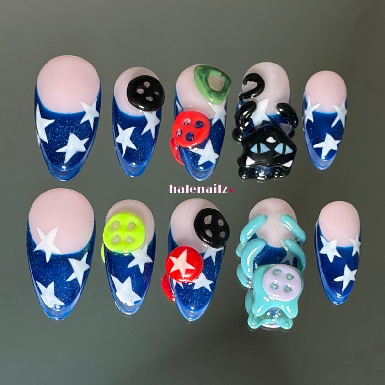 May include: A set of ten press-on nails featuring a dark blue base with white star patterns and a nude-colored cuticle area. The nails are decorated with various three-dimensional elements including black, red, and neon green buttons and cat figures.