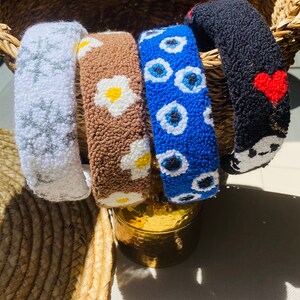 May include: Four colorful headbands with different patterns. The headbands are made of a soft, fuzzy material. The patterns include white stars, brown and white daisies, blue evil eyes, and black with a red heart.