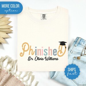 May include: Cream-colored t-shirt with the word "Phinished" in pastel colors and a graduation cap graphic. The text "Dr. Olivia Williams" is below the word. The shirt is folded on a white surface with denim jeans.